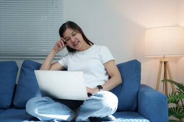 Wall Mural - Young asian woman using laptop computer working from home sitting on blue sofa in living room, studying online, watching webinar, making video call, freelancer writing blog