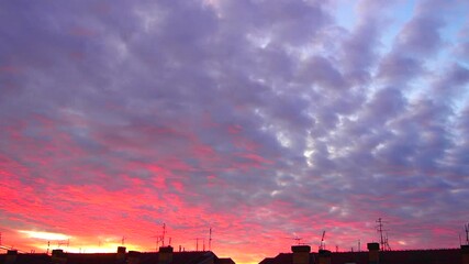 Poster - Bright pink hues dominate the sky as the sun sets, casting a vibrant glow over rooftops in the evening. Clouds reflect the warm colors, creating a serene atmosphere. Real time. Dramatic cumulus clouds