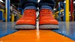 © NAPHAT - Safety First: A close-up view of a pair of orange safety shoes with black laces, standing on a blue and orange striped floor in an industrial setting. The shoes are worn by a person in blue jeans.
