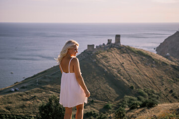 Wall Mural - A blonde woman stands on a hill overlooking the ocean. She is wearing a white dress and she is enjoying the view.