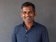 © ANUAR - Friendly South Asian man smiling in a casual blue polka dot shirt against a neutral gray background, exuding warmth and approachable demeanor for lifestyle imagery.