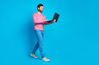 © deagreez - Confident young man in casual attire walking while using laptop on vibrant blue background, portraying modern technology and digital lifestyle