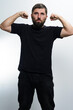 © Niko_Dali - A bearded man in a black t-shirt and dark jeans flexes his biceps against a light gray backdrop, conveying strength, confidence, or pride.