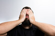 © Niko_Dali - Close-up of a man with a beard covering his face with his hands against a plain background. He appears distressed, ashamed, or experiencing a negative emotion.