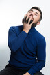 © Niko_Dali - A bearded man wearing a blue turtleneck sweater and dark pants sits, thoughtfully stroking his beard and looking upwards against a white background.