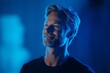 © Maelgoa - A tall, lean man in his late 30s with blond hair and a beard, standing confidently in a blue-lit void, his relaxed expression and smile illuminated by soft lighting 1