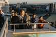 © qunica.com - A diverse group of business colleagues brainstorm project solutions while standing on a high tower balcony, demonstrating teamwork and innovative thinking during a sunset meeting in an urban setting.