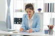 © Nittaya - Focused Businesswoman Analyzing Data: A young, Asian businesswoman in a light blue blazer intently reviews financial documents at her modern, minimalist desk.