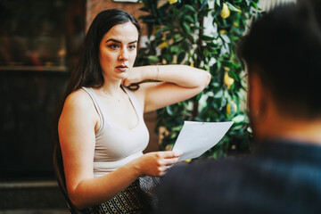 Wall Mural - A young woman attentively discusses ideas with a colleague in a trendy coffee shop. Holding a paper, she appears focused and engaged, embodying collaboration and creativity in a professional setting.