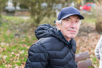 An elderly man in a jacket and hat sits alone on a bench in a park. The concept of loneliness in old age