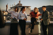 © qunica.com - A group of multiracial business colleagues relaxes on a rooftop balcony, celebrating a successful day as the sun sets over the cityscape, showcasing camaraderie and diversity in a professional setting