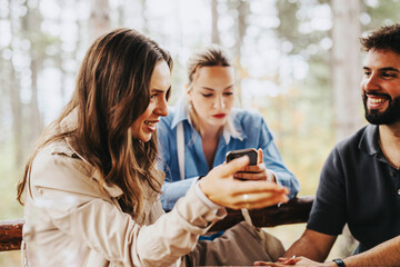 Poster - A group of friends engaging in lively conversation outdoors. They are smiling and using a smart phone, surrounded by a serene forest background, creating a warm and joyful atmosphere.