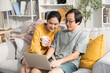 © saksit - Asian couple relaxing on sofa at home and using laptop