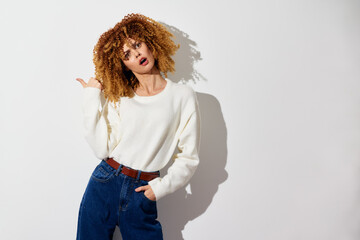 Wall Mural - Woman with curly hair posing confidently against a white background, wearing a comfortable cream sweater and blue high waisted jeans, showcasing a stylish, casual look