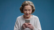 ©  Mohammad Xte - Elderly woman with short wavy hair, wearing a light-colored sweater, smiling as she looks at a smartphone in her hands. The solid blue background highlights her cheerful expression