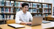 © sevenSkies - Asian young man typing on laptop at desk in library wearing white shirt