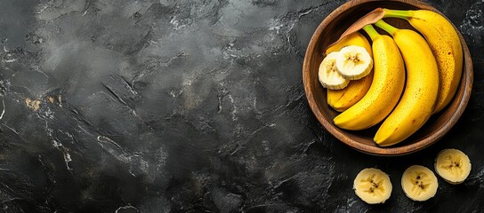 Wall Mural - Ripe yellow bananas in a wooden bowl, sliced bananas on dark background.