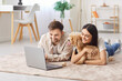 © Studio Romantic - Young family couple resting together with their maltipoo dog, lying on carpet on floor at home. Smiling woman hugging puppy, watching funny video or film on laptop with her joyful husband and pet.