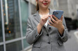 © Parichat - Businesswoman wearing gray blazer and white shirt, using smartphone outdoors, browsing internet and social media, standing in front of modern office building