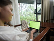 © SHOTPRIME STUDIO - A young woman in casual white clothing sitting comfortably at home, using a laptop with a green screen, surrounded by greenery and natural light, representing modern remote work