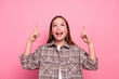© deagreez - Cheerful preteen girl wearing checkered shirt expressing excitement with fingers pointing upwards against vibrant pink background.