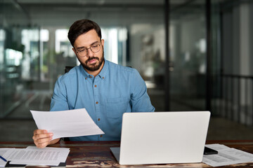 Busy Latin financial expert checking documents working at laptop in office. Serious business man accountant reading law paper company file invoice overview looking at computer at workplace.