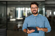 © insta_photos - Portrait of smiling male company worker employee, project manager or entrepreneur in office. Happy young professional Latin business man looking at camera standing at work using tablet.