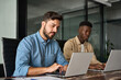 © insta_photos - Two busy diverse male employees using laptop computer talking in office. Professional young business men workers team having conversation working on project sharing ideas sitting at work desk.