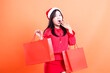 © Ajab - Asian young female expression wearing red blouse and santa hat, standing sideways to left surprised to camera hand lifting red bag of christmas gifts and covering mouth, isolated on orange background