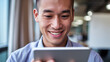 © Tom - Smiling young Asian man engaged with his tablet in a modern office setting, radiating positivity and focus.