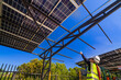 © ultramansk - A female engineer in a safety vest and helmet inspecting a solar carport under construction. The structure’s steel beams and partially installed solar panels are visible under a bright blue sky.