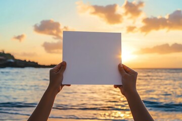 Wall Mural - a person holds a blank sheet of paper against a sunset by the beach.