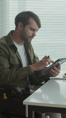 Vertical shot of Caucasian businessman in wheelchair using digital tablet while working at office desk