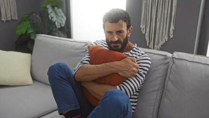 Wall Mural - Young man with beard hugging pillow on a couch feeling upset in living room with plants