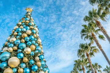 Wall Mural - Festive Christmas tree adorned with blue and gold ornaments against a vibrant blue sky and palm trees. AI.