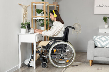 Wall Mural - Young woman in wheelchair with headphones using laptop on table at home