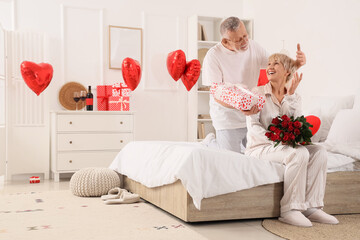 Poster - Mature man greeting his happy wife with gift box and bouquet of roses on bed at home. Valentine's Day celebration