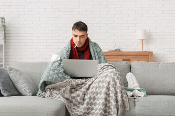 Wall Mural - Ill young man with thermometer using laptop on sofa at home