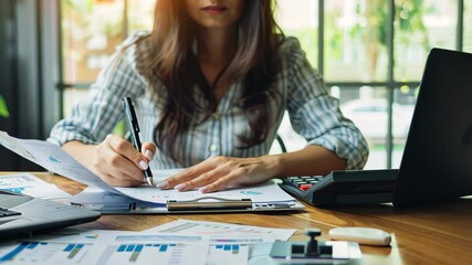 Poster - A woman is focused on her work at her desk, reviewing financial documents while using a laptop and writing notes, surrounded by a bright office environment.