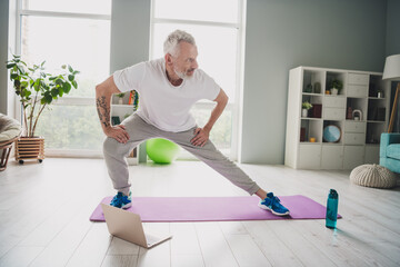 Wall Mural - Elderly man doing stretching exercises on yoga mat at home in bright room with laptop nearby, embracing a healthy lifestyle and staying fit