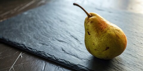 Aufkleber - A single ripe pear, displaying its golden skin and delicate stem, rests gracefully on a smooth, dark slate surface.