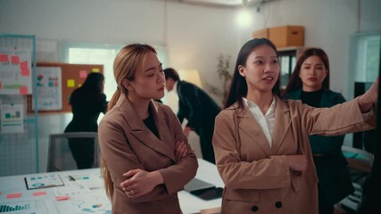 Fototapete - Professional asian businesswomen collaborating during strategic meeting, pointing at data visualizations while colleagues work in contemporary corporate environment