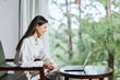 © SHOTPRIME STUDIO - Young woman in casual white shirt sitting by a laptop, engaged in remote work, showcasing focus and serenity against a green nature backdrop