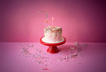 Poster - A pink and white birthday cake with a single lit candle sits on a red cake stand in front of a pink background. Sprinkles are scattered around the cake.