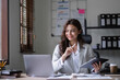 © wichayada - Young Businesswoman Analyzing Documents at Desk in Modern Office Setting with Laptop and Tablet