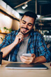 © BullRun - Smiling hipster guy reading new message in social networks confirming new friend on smartphone, positive young man satisfied with banking app on mobile phone checking balance while learning