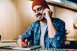 © BullRun - Cheerful male creative designer drawing sketch in notebook having telephone conversation, smiling hipster guy making telephone call to friend for discussing ideas while sitting in college library