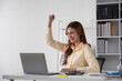 © wichayada - Young Businesswoman Celebrating Success at Work, Happy and Excited, Sitting at Desk with Laptop in Modern Office
