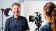 © Vilaysack - A smiling man stands in front of a camera, engaged in a video shoot with a soft-focus woman behind the camera in a well-lit studio.