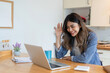© wichayada - Young Freelance Woman Working from Home in Dining Room, Smiling and Waving at Laptop Screen, Remote Work Concept